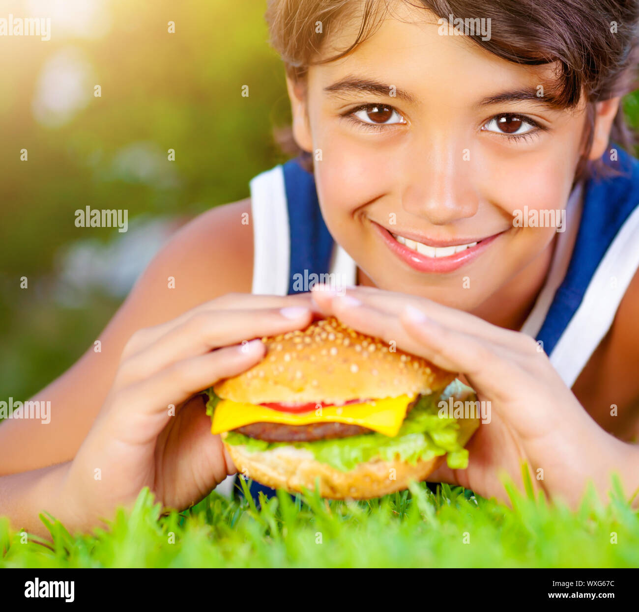 Closeup portrait of cute happy boy eating big tasty fatty burger outdoors, lying down on green field and enjoying sandwich with cheese, meat and veget Stock Photo