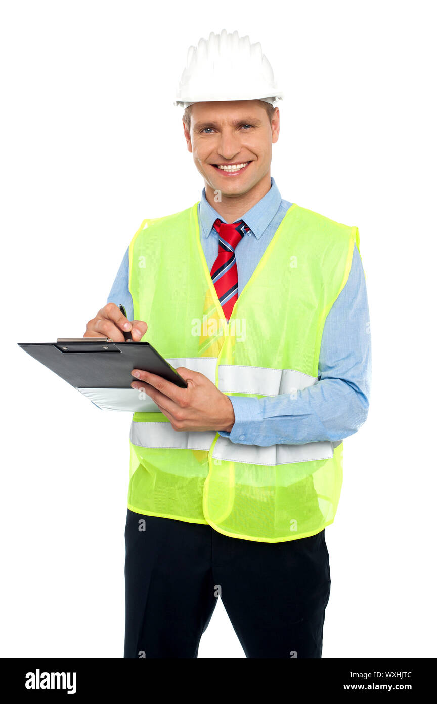 Smiling young construction engineer with safety hat writing on notepad against white background Stock Photo
