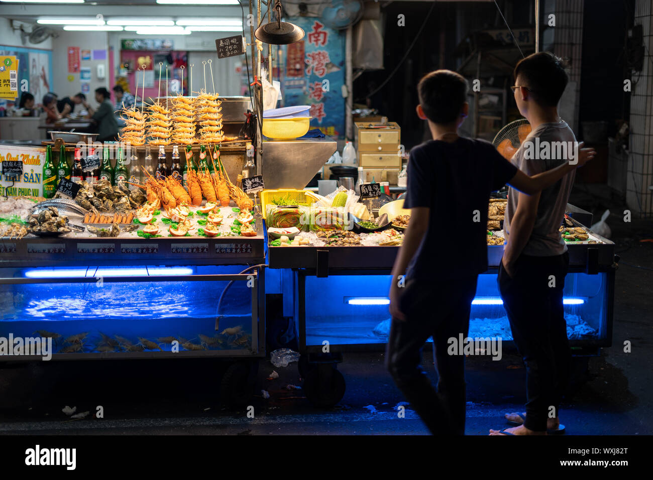 Kaohsiung, Taiwan: Two boys standing in front of a Seafood street food stall at the LiuHe Nightmarket (六合夜市) thinking about what to buy. Stock Photo