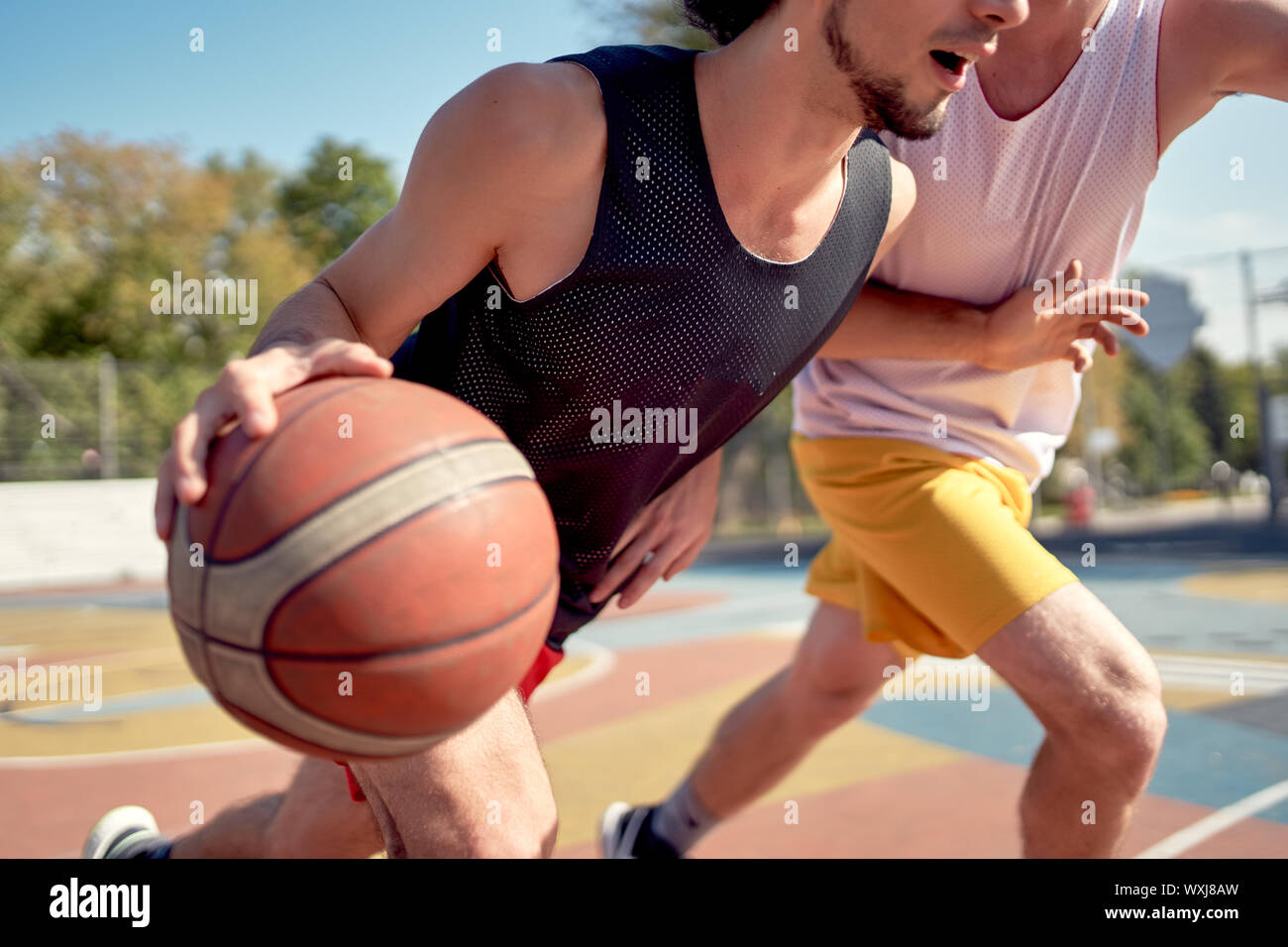 Photo of young athletes playing basketball on playground on summer day against background of green trees Stock Photo