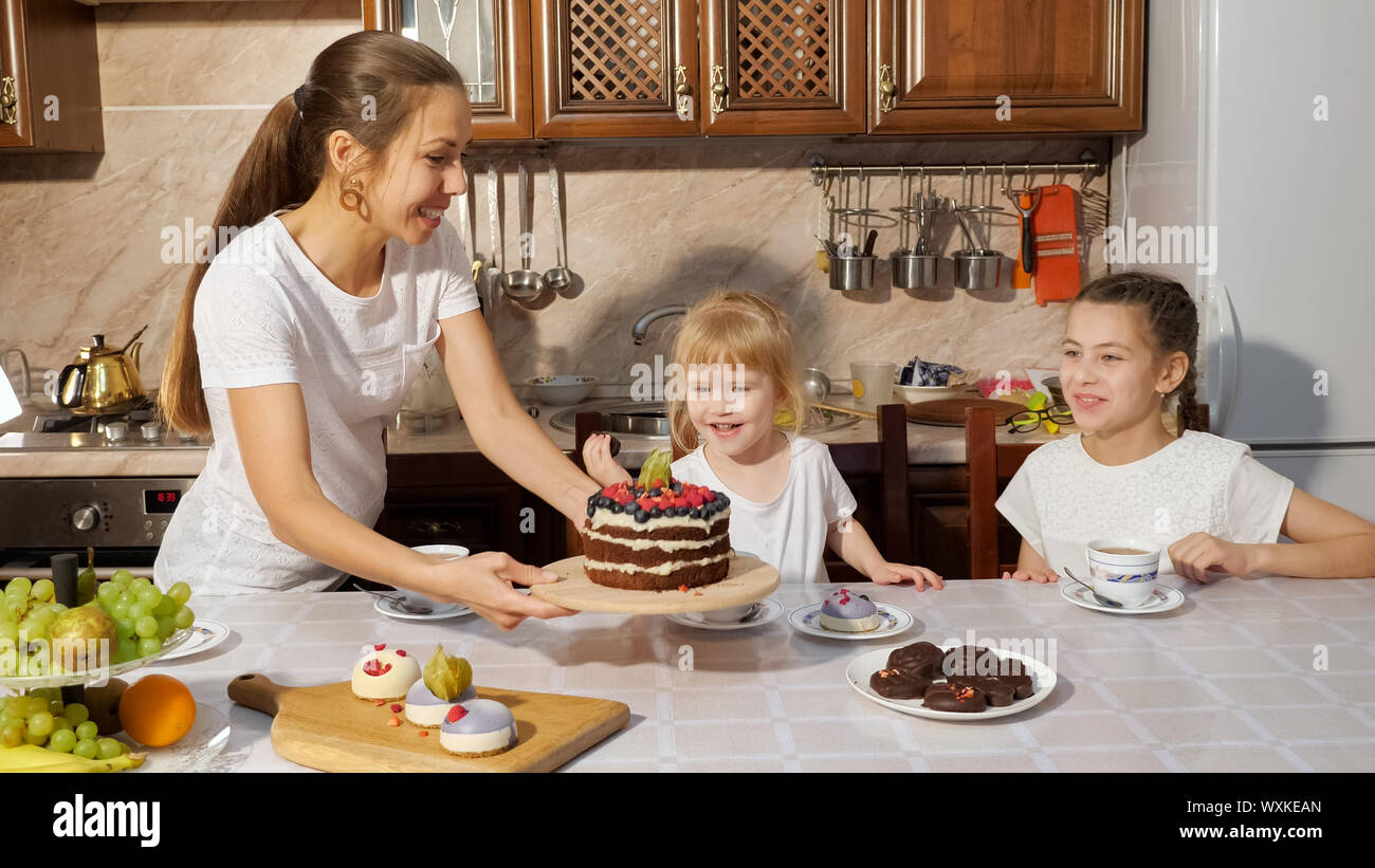 Birthday surprise from mom. Mother brings the chocolate homemade cake to her daughters having a tea sitting in kitchen at home. Stock Photo