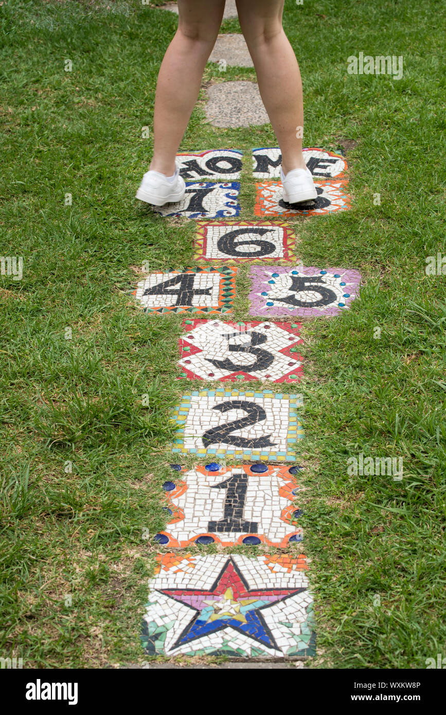 A young girl in white sneakers plays hopscotch on colourful glazed tiles embedded in a lawn in Australia Stock Photo