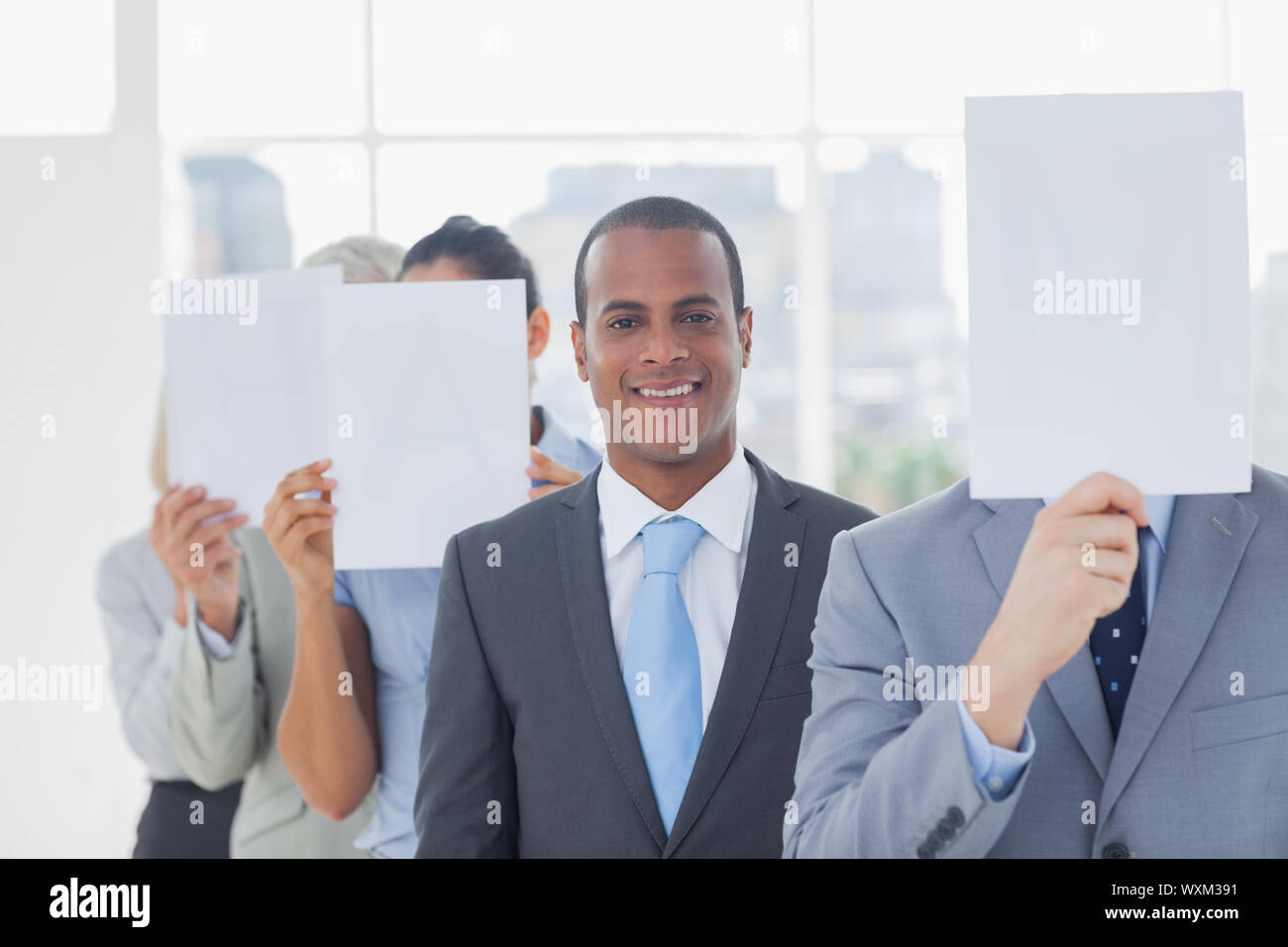 Businessman smiling at camera with colleagues covering faces with white pages Stock Photo