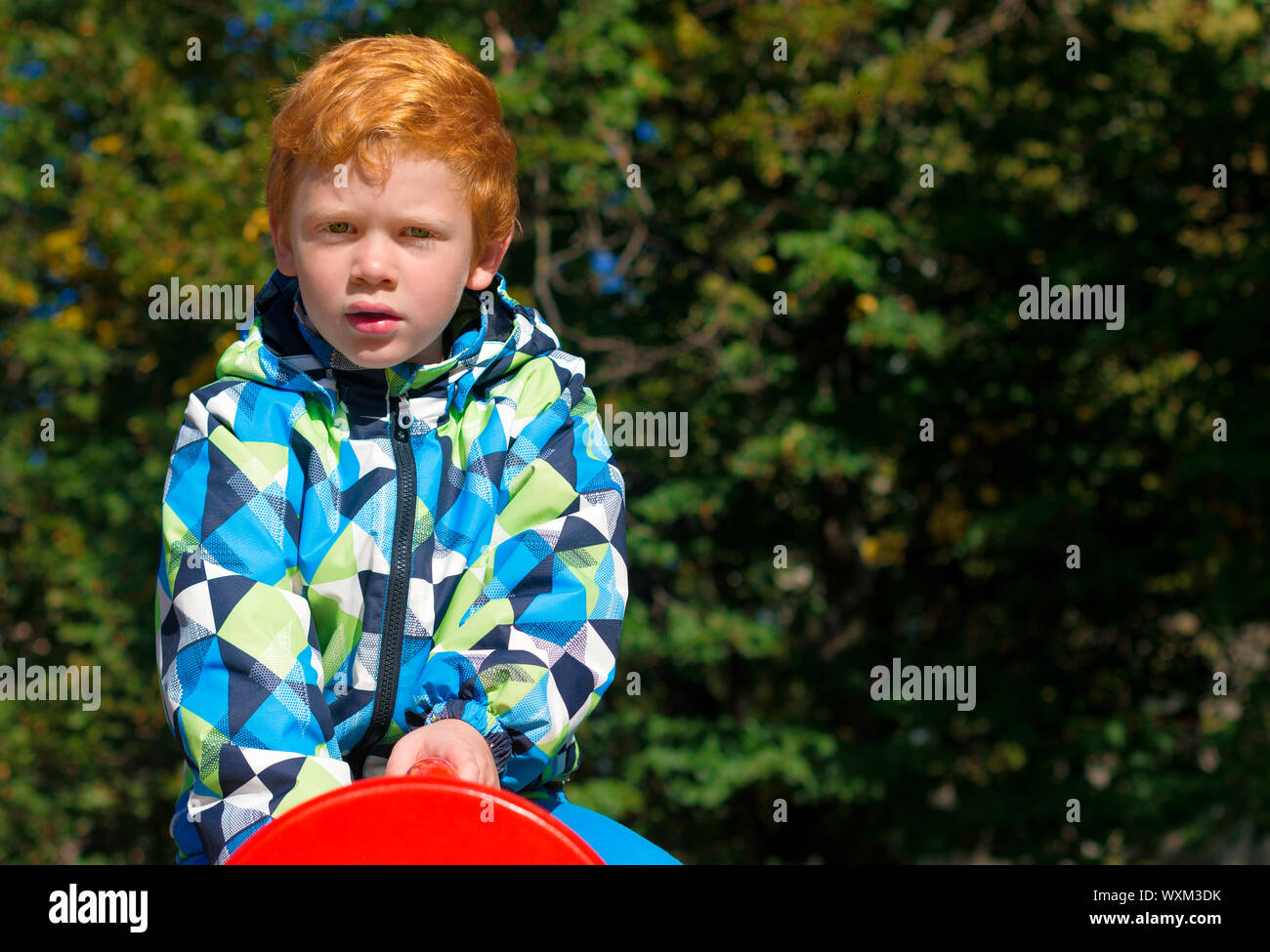 Child playing on outdoor playground. Boy play on school or kindergarten yard. Kid with curly ginger hair. Portrait. The red-haired boy stares intently Stock Photo