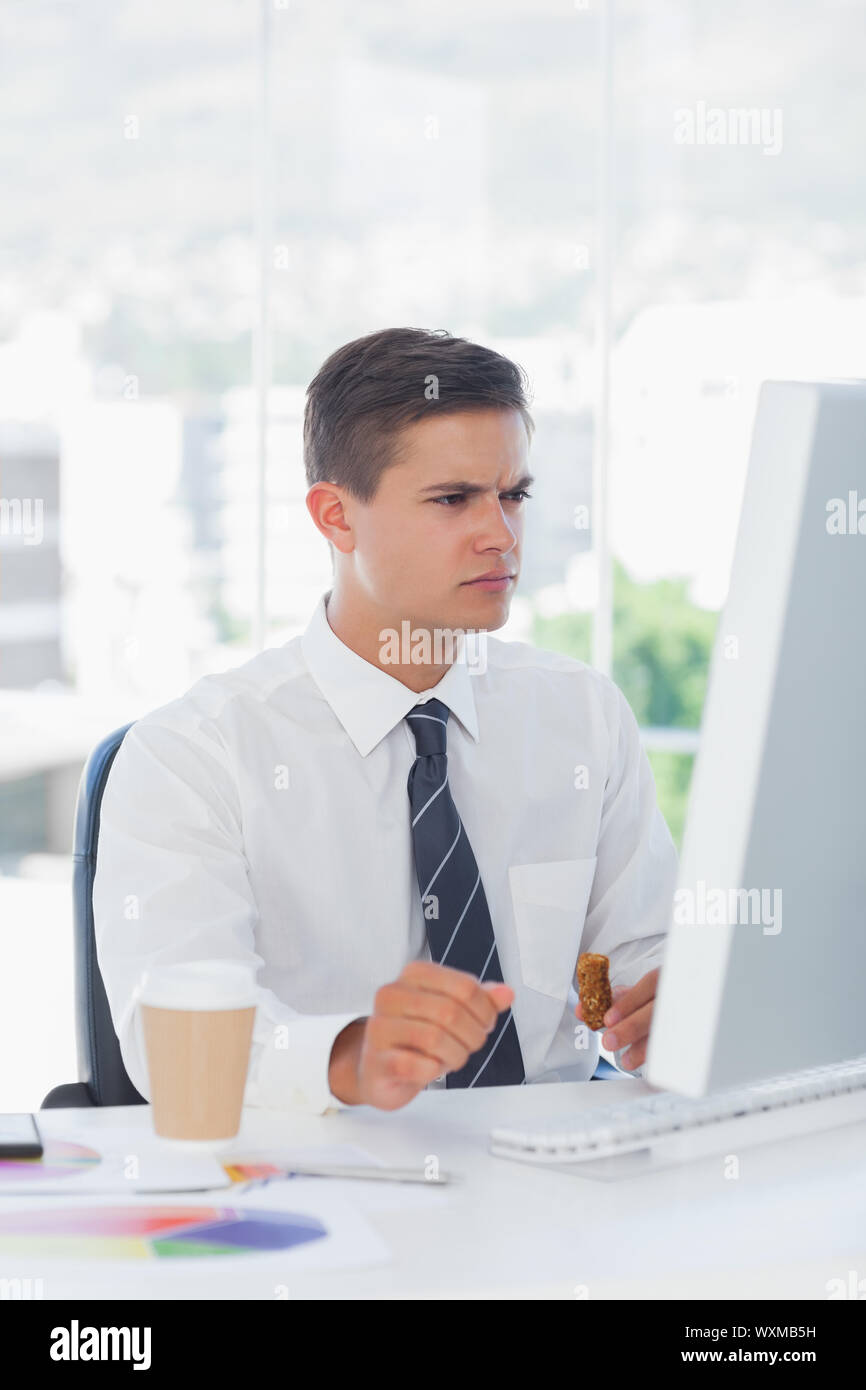 Young businessman frowning while looking at computer and eating Stock Photo