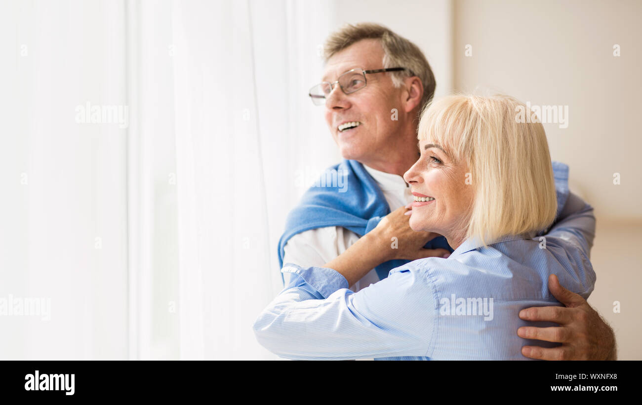 Happy senior couple looking through window and hugging Stock Photo