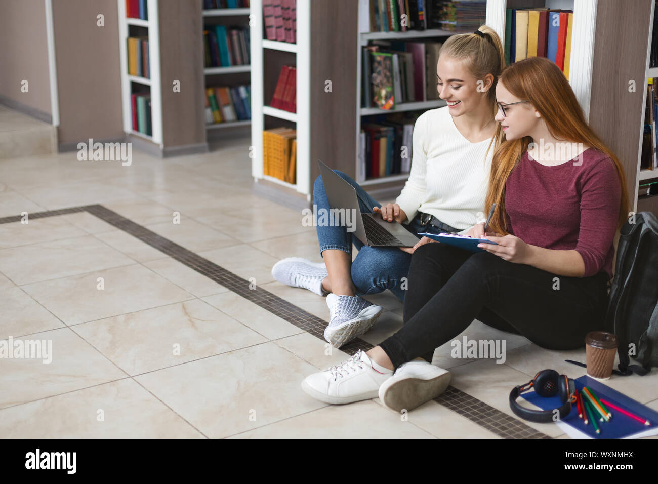 Two girlfriends sitting on floor at library, studying with laptop Stock Photo