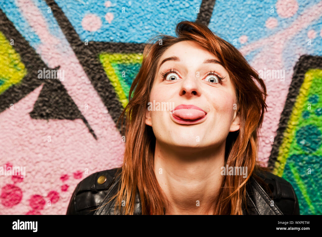Stylish girl poking out her tongue to the camera against colorful graffiti wall. Stock Photo