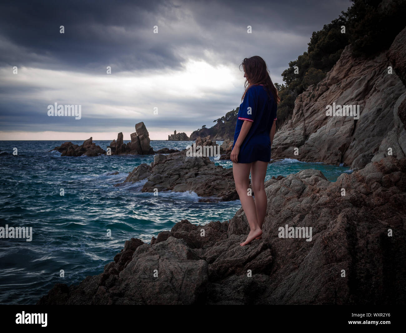 Caucasian brunette girl standing on the rocks near the rippling sea under dramatic skies in the twilight Stock Photo