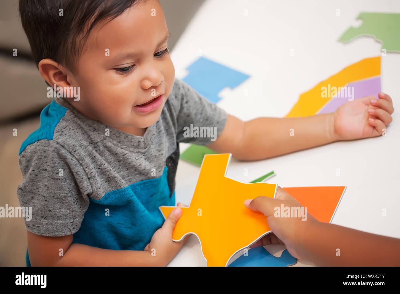 Two kids working together to assemble a North American puzzle, where one child hands over the shape of the state of Texas to a boy. Stock Photo