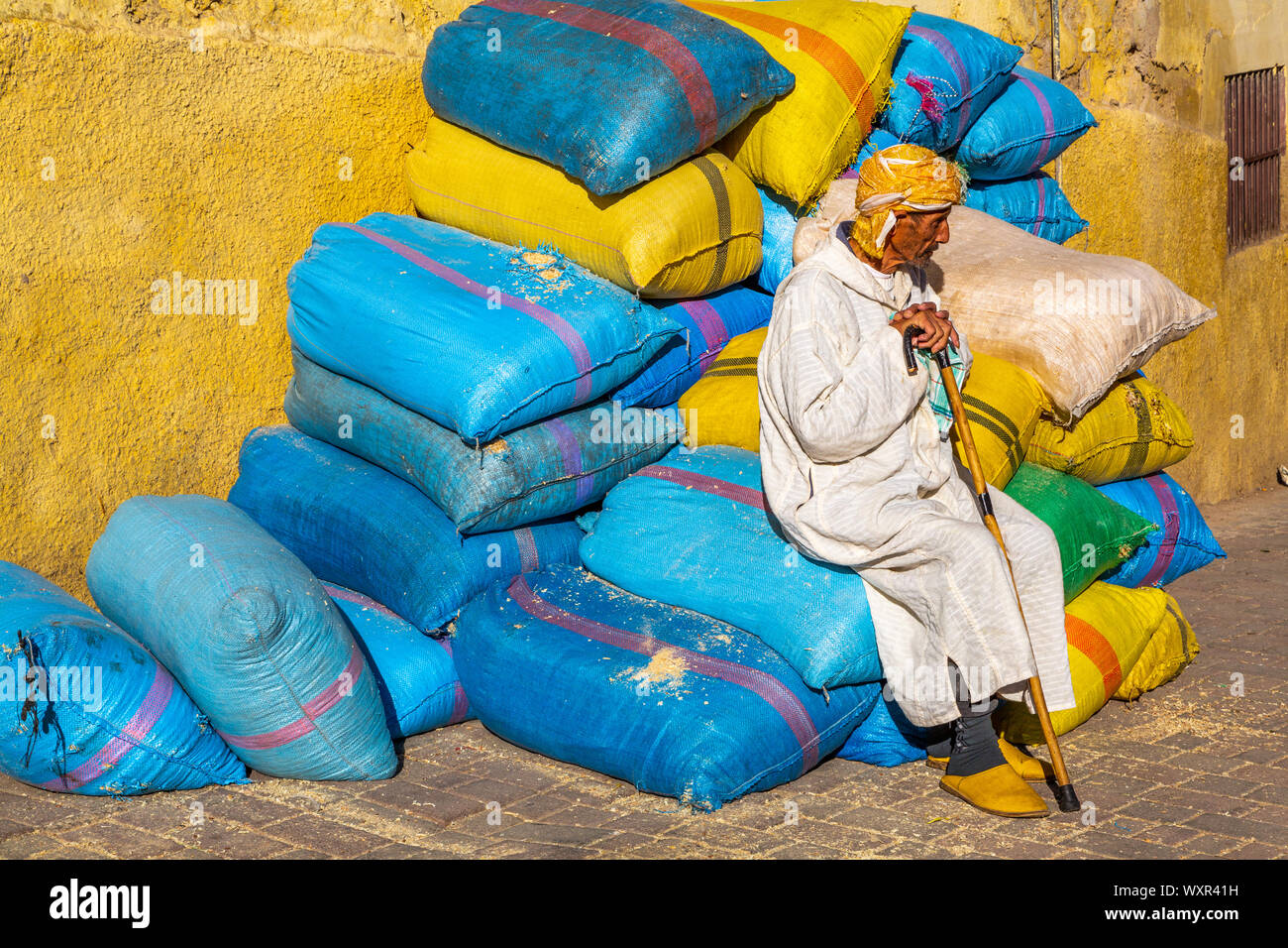 Elderly traditionally dressed Moroccan man with walking stick sitting on colourful plastic bales of food ingredients by yellow wall, Marrakech, Morocco Stock Photo