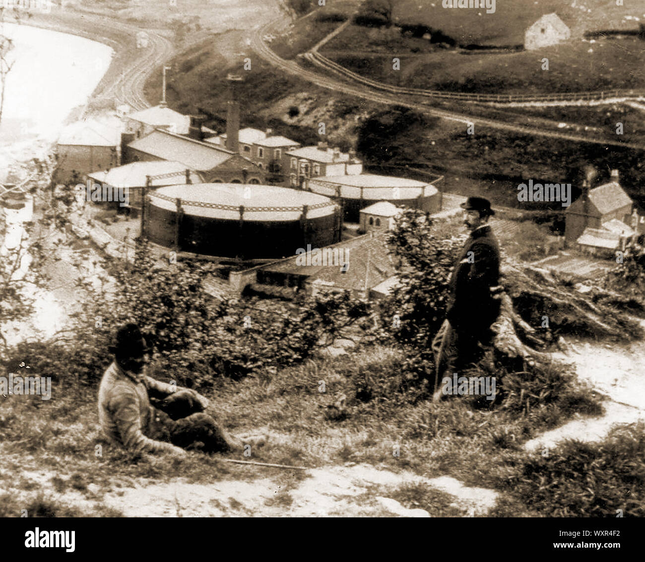 A Victorian picture of the old gas works (now demolished) at Whityby North Yorkshire, England. The picture is taken from the East bank of the River Esk which would later be connected to the east side by a railway viaduct to be built over the gasworks site. The railway lines from Whitby to Middlesbrough via the coast, and Whitby to Middlesbrough via the Esk Valley can be seen opposite the two men. The derelict gasworks site was later reclaimed by  Fif Robinson who lived there using home-made solar panels to provide electricity. Stock Photo