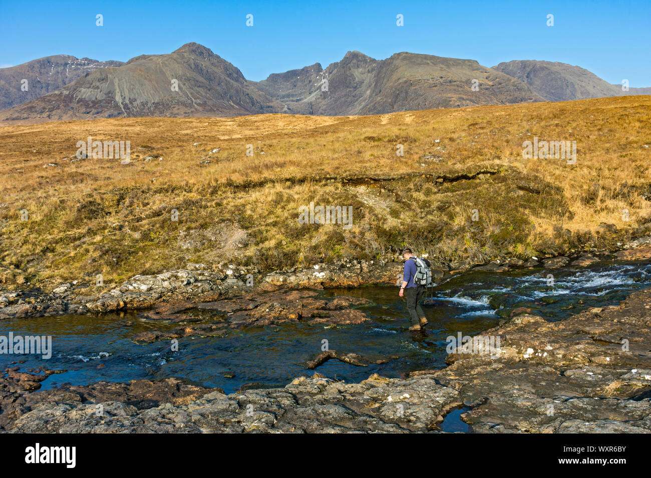 Walker crossing the Allt Coire Lagan stream with the Cuillin mountains behind, on the track to Rubha an Dùnain, Minginish, Isle of Skye, Scotland, UK Stock Photo