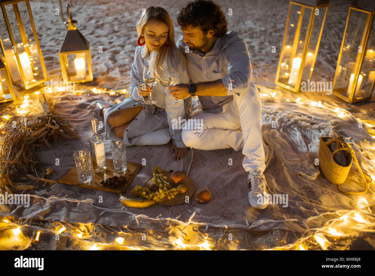 Couple having a romantic dinner, hugging and clinking wine glasses at the beautifully decorated place illuminated with different lights the sandy beach at dusk Stock Photo