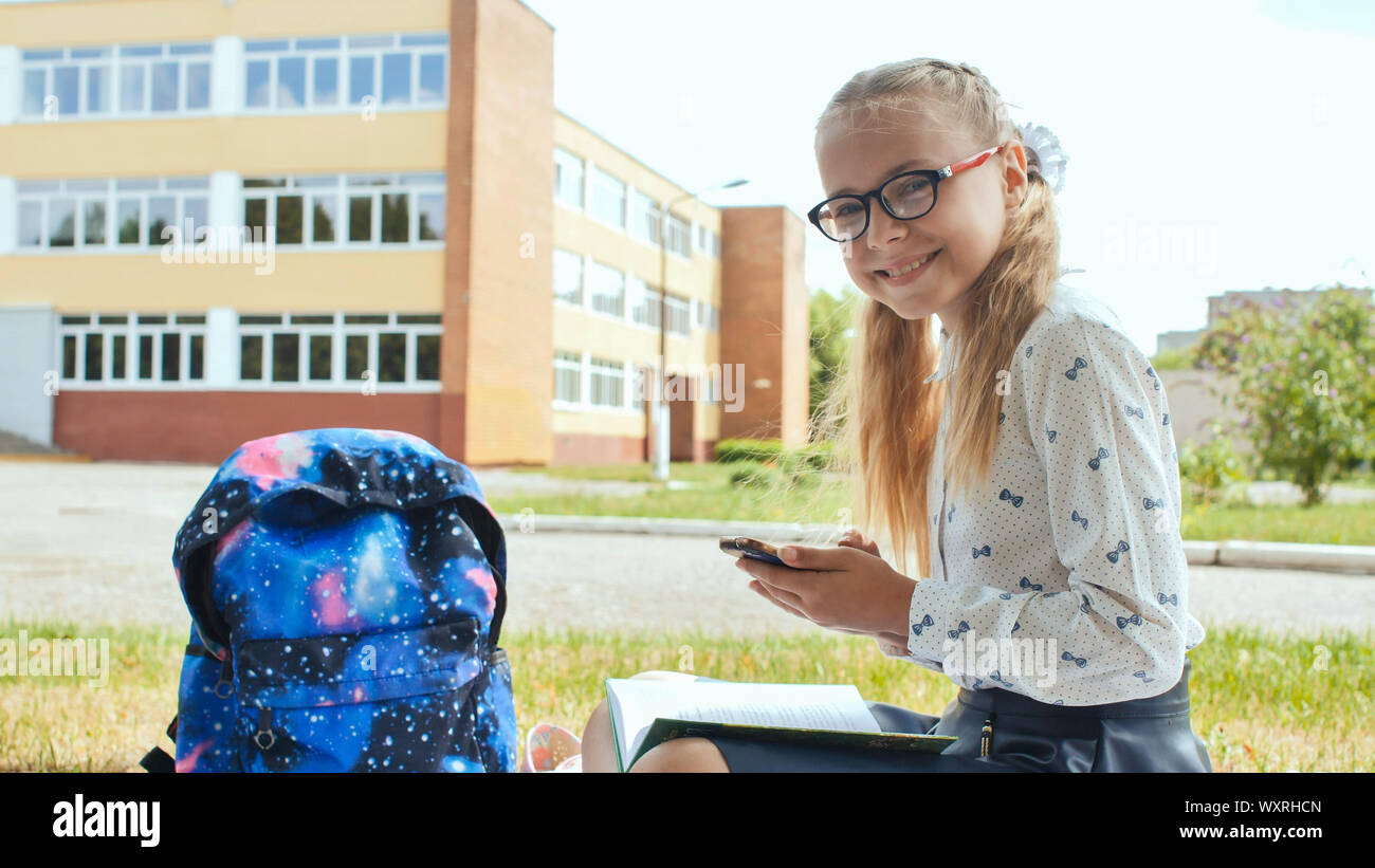 An eleven-year-old schoolgirl sits on the grass with a book and looks into the phone. Stock Photo