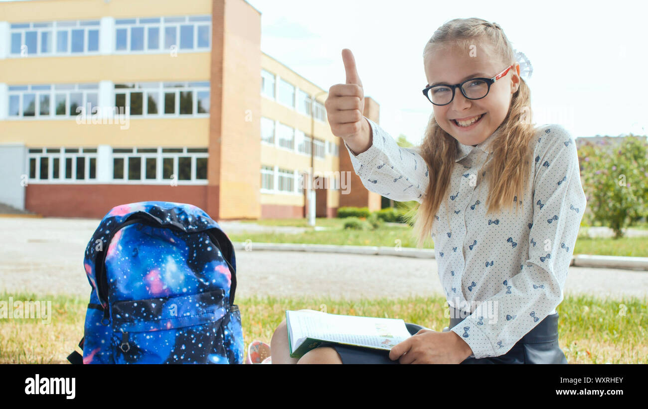 An eleven-year schoolgirl with a book sits on the grass and shows a finger in up. Good mood. Stock Photo