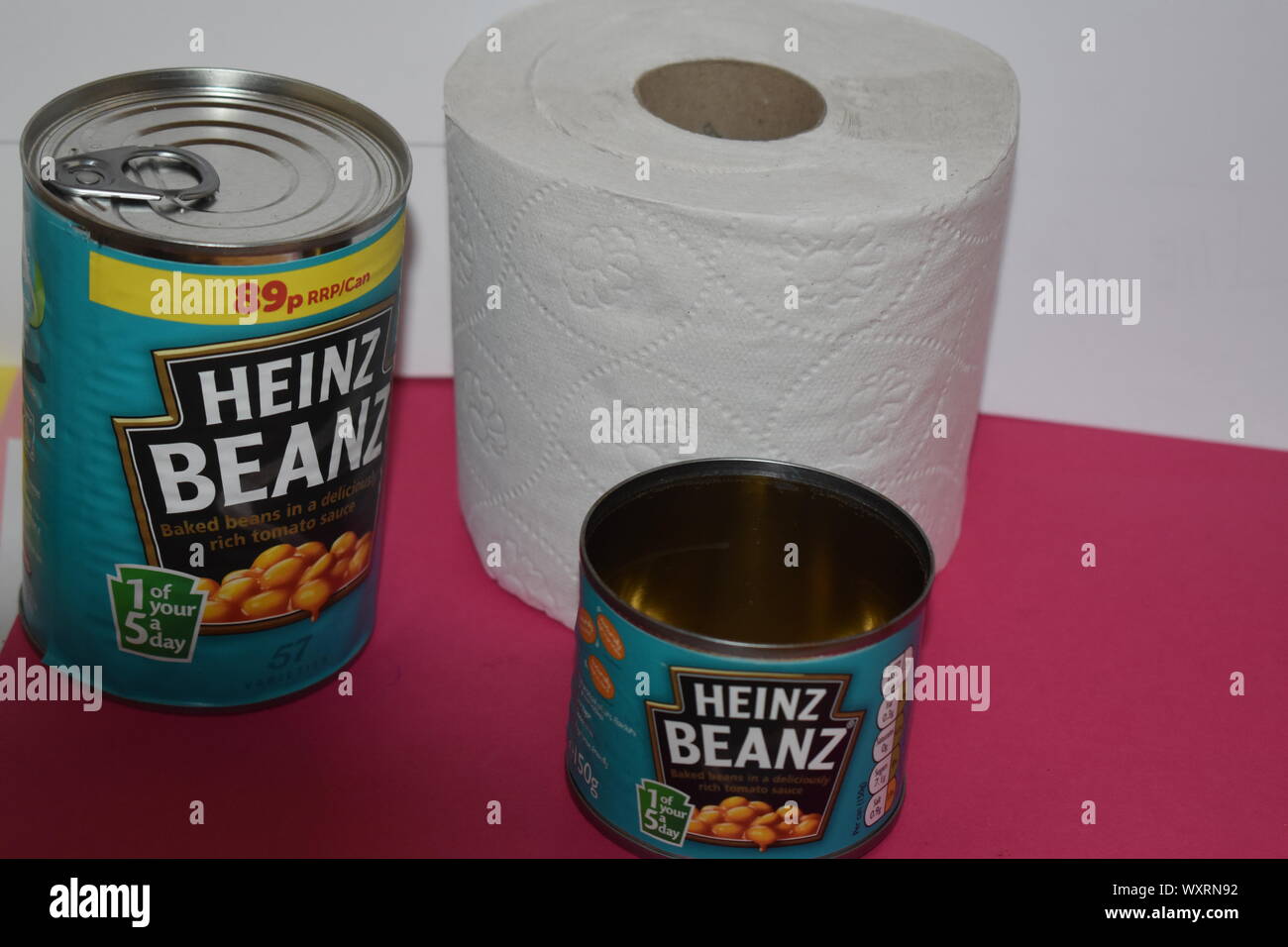 Two tins of Heinz baked beans. 1 large, 1 small with effect on dark pink and white background and a roll of bog standard toilet tissue. Fibre farts. Stock Photo