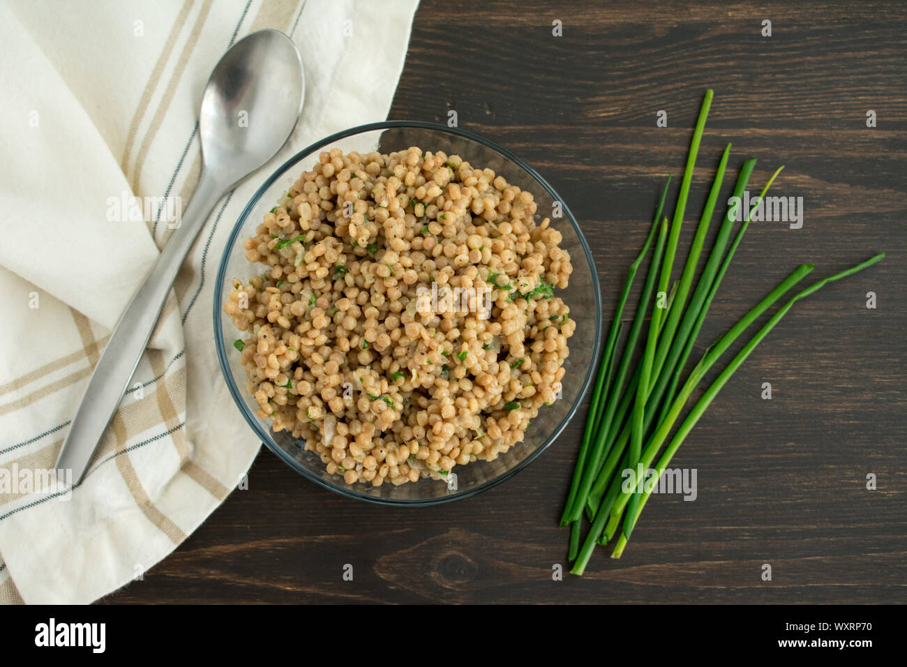 Herbed Couscous Pilaf with Chives: A bowl of whole wheat pearl couscous with fresh chives, a spoon, and a napkin. Stock Photo