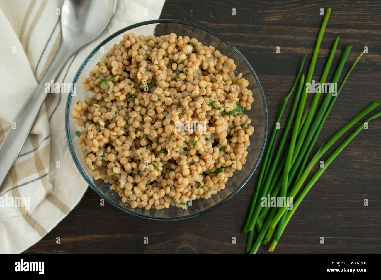 Herbed Couscous Pilaf with Chives: A bowl of whole wheat pearl couscous with fresh chives, a spoon, and a napkin. Stock Photo