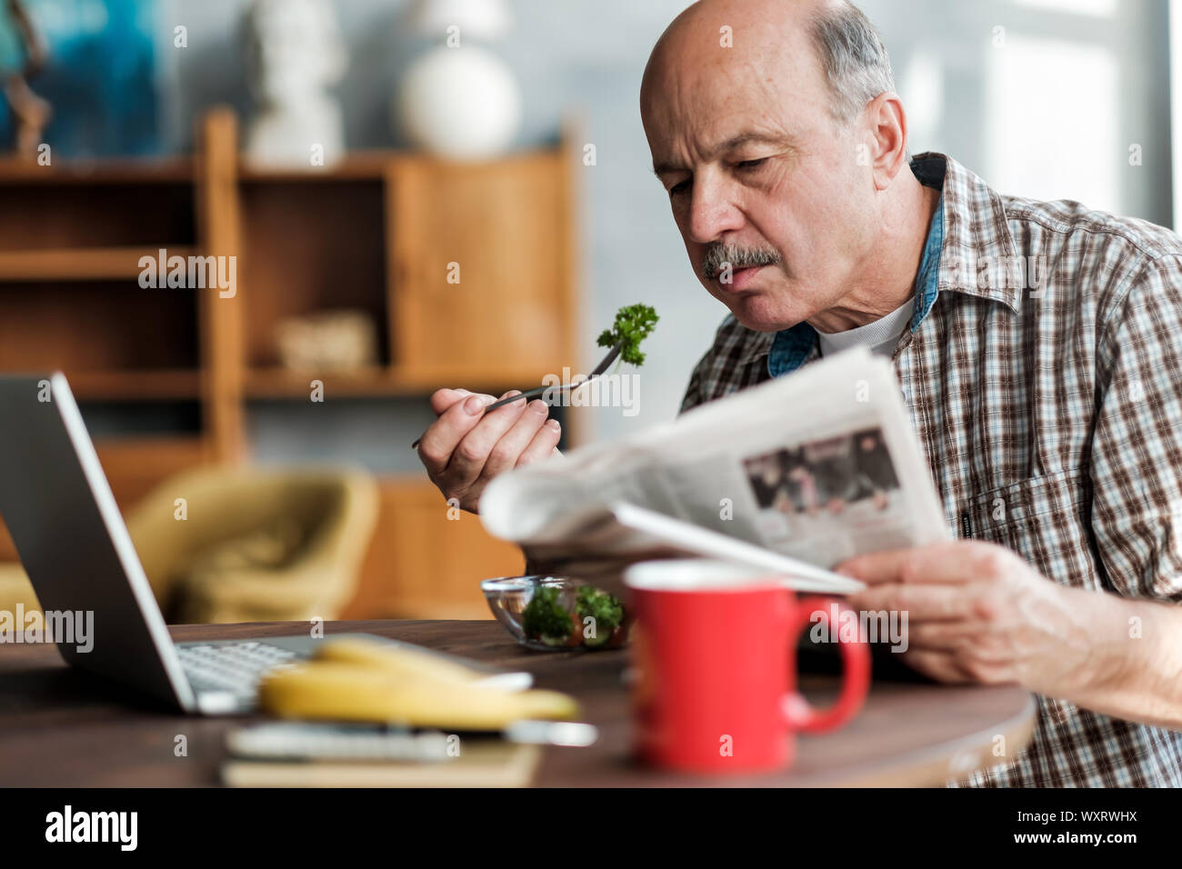 Senior hispanic man eating fresh salad in the morning sitting in the living room near laptop Stock Photo