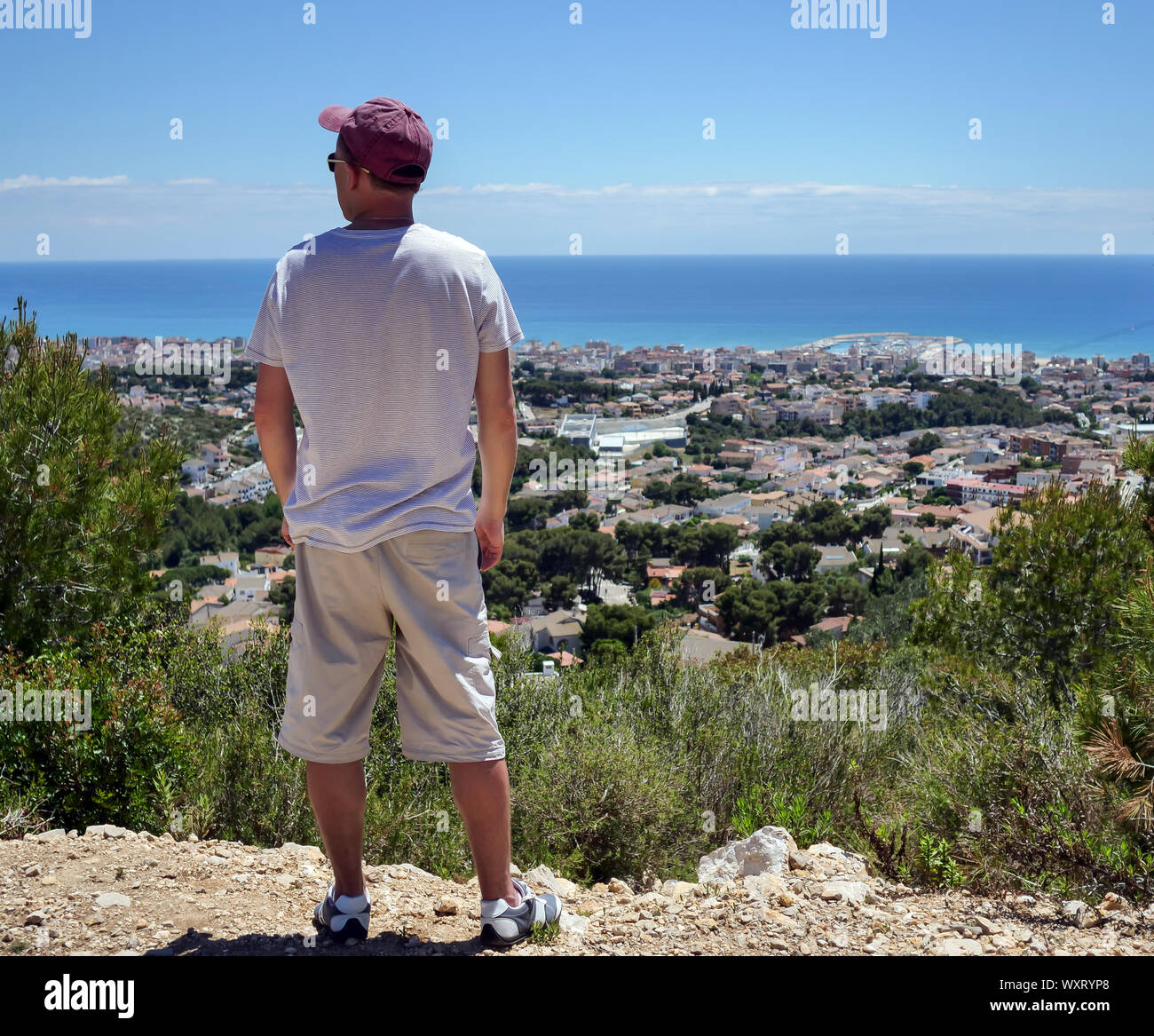 Young guy tourist stands on a mountain and looks at the city near the sea at the foot of the mountain, vacation concept Stock Photo