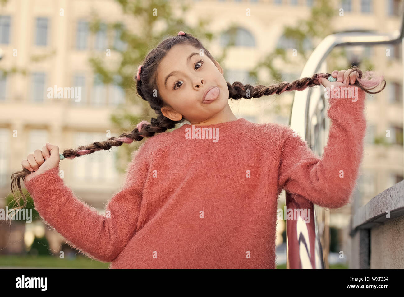 Having fun with her hair. Funny girl holding hair plaits on urban background. Small cute girl with long brunette hair showing tongue outdoor. Little hair model with crazy look Stock Photo - Having fun with her hair. Funny girl holding hair plaits on urban background. Small cute girl with long brunette hair showing tongue outdoor. Little hair model with crazy look Stock Photo -