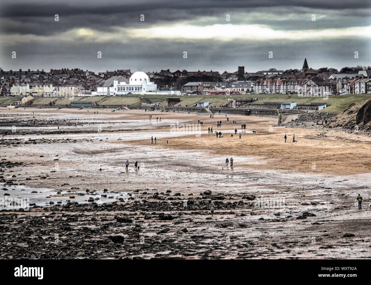 Strollers along the beach at Whitley Bay, in the north of England on a dreary winter afternoon with the white outline of the Spanish City illuminated Stock Photo