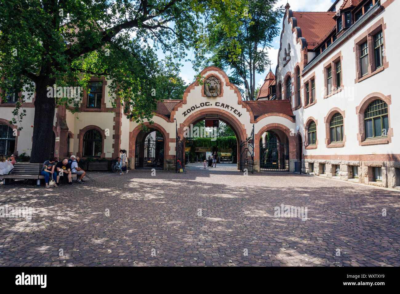 Leipzig German July 09 2018: The open gate of the Leipzig Zoo with a father and child entering while beside the entrance on a bench people in the sun Stock Photo