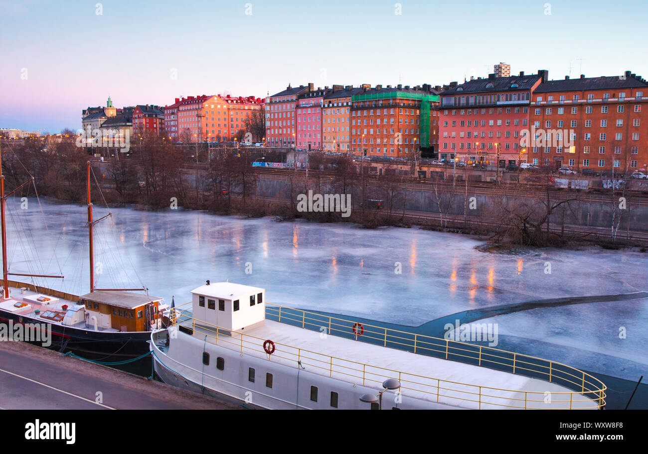 Boats frozen in the ice of Karlberg Lake (Karlbergssjon) at dawn with colourful facades of Atlas district houses, Norrmalm, Stockholm, Sweden Stock Photo
