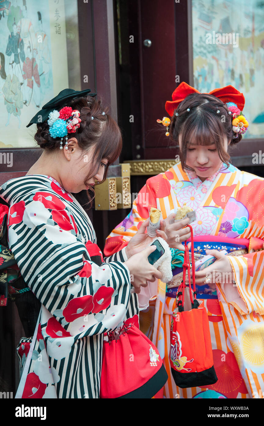 Two young Asian women in richly coloured kimonos look at mobile phones in front of building. Springtime, cherry blossom season, Tokyo, Japan Stock Photo