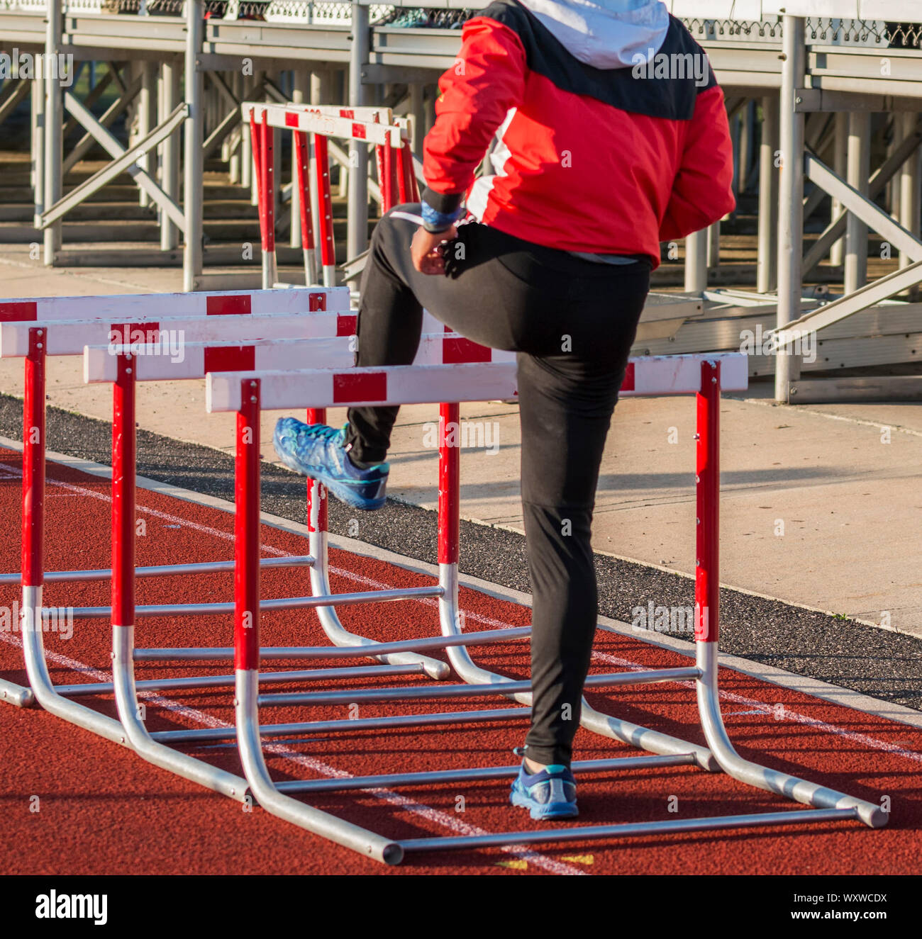 A high school girl track and field athlete is warming up for her hurdle race by walking over hurdles on a red track in her sweatsuit. Stock Photo