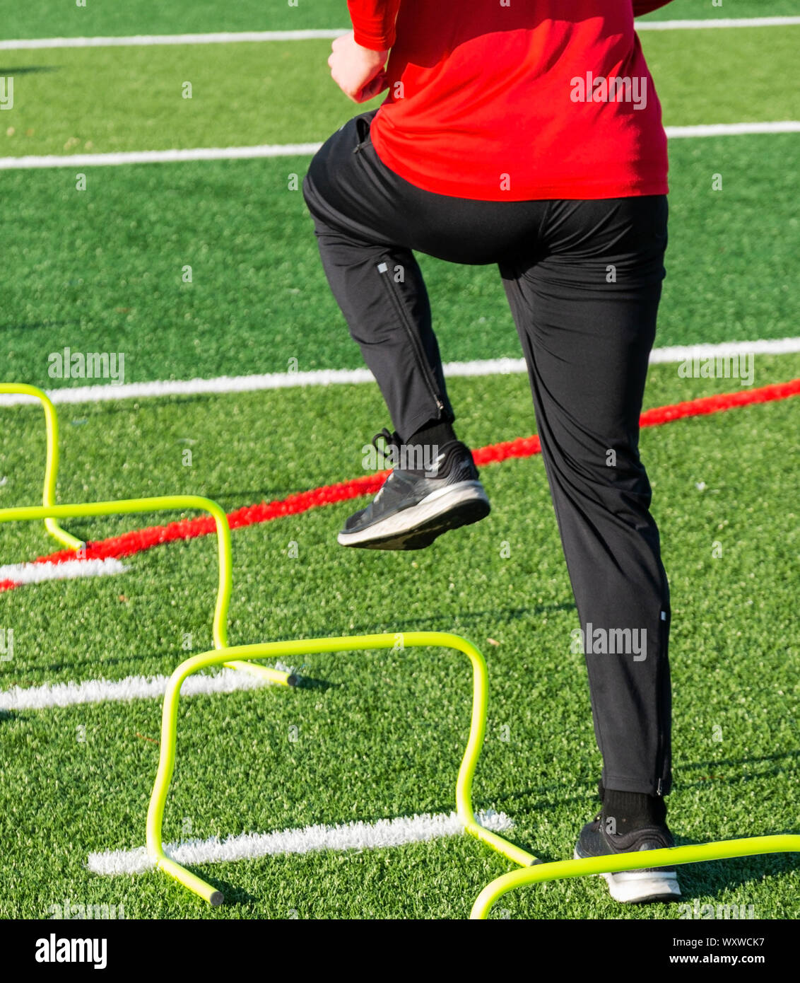 A high school athlete is performing speed and agility sports training drills stepping over yellow mini hurdles on a green turf field. Stock Photo