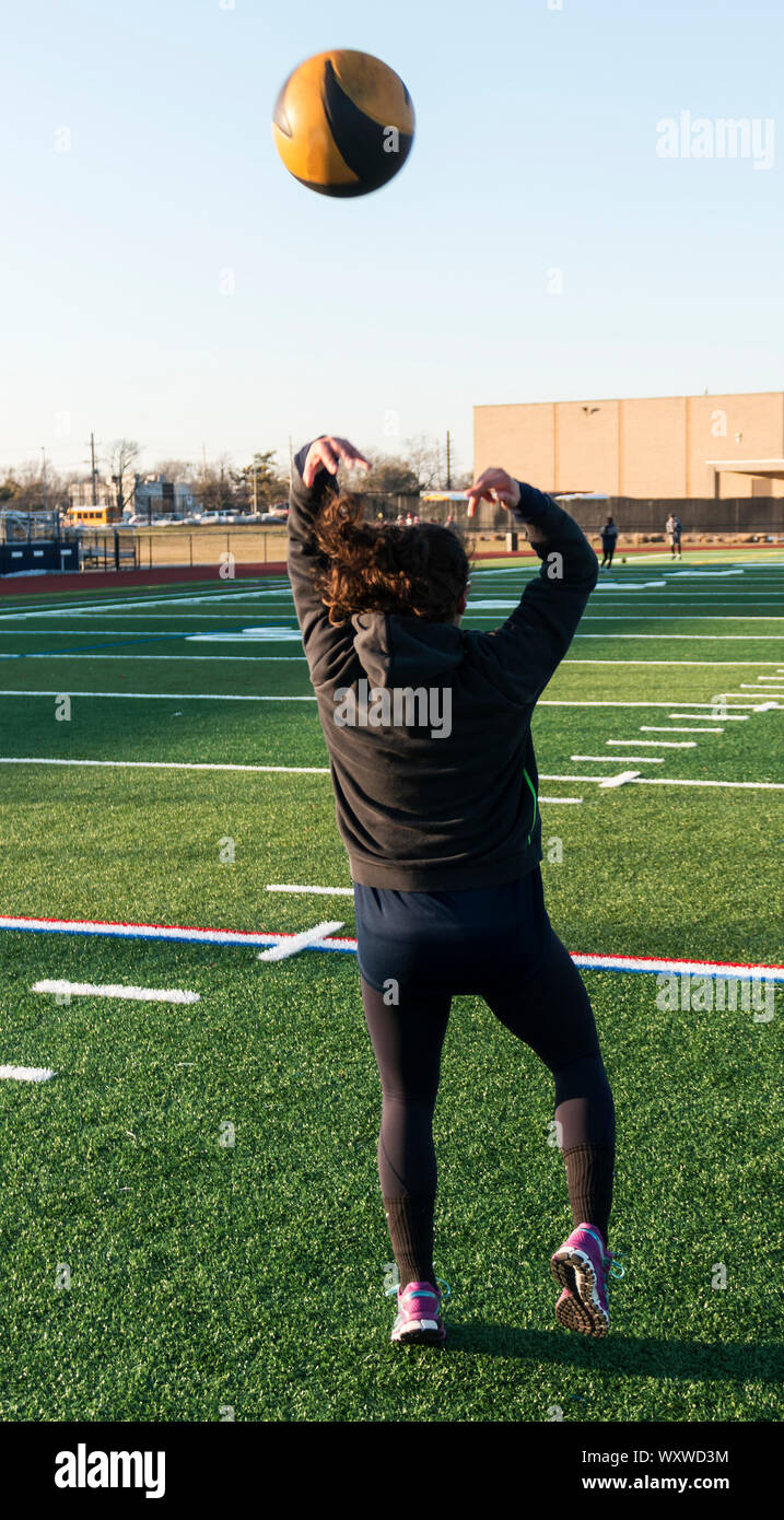 An athlete is on a green turf field throwing a black and gold medicine ball over her head backwards during track and field practice. Stock Photo