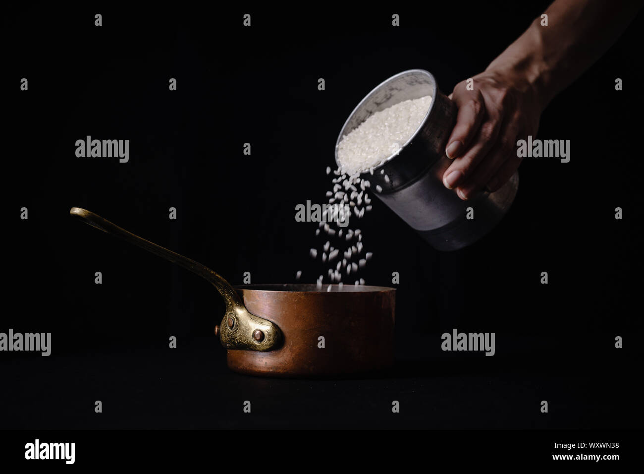 Female hand pouring arborio rice to copper saucepan for boiling. Dark background, copy space
