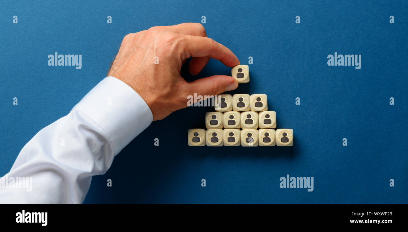 Male hand stacking wooden dices with person icons on them in a pyramids shape in a conceptual image of business hierarchy. Over navy blue background. Stock Photo