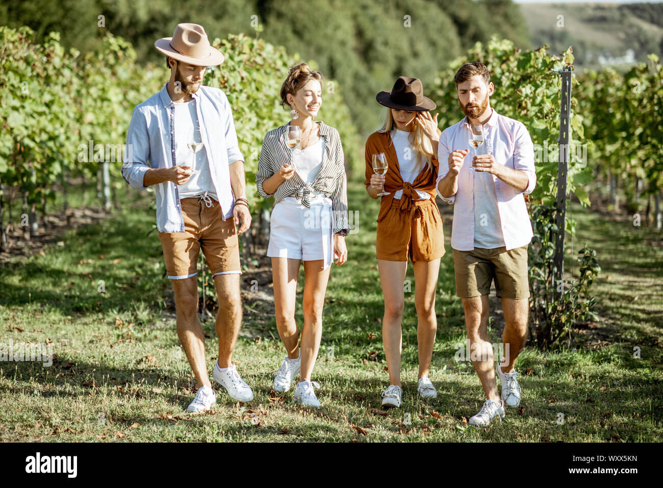 Group of young friends dressed casually having fun together, walking with wine glasses on the vineyard on a sunny summer morning Stock Photo