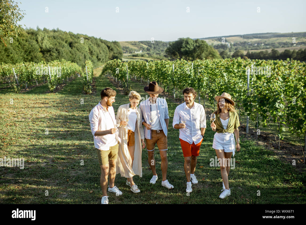 Group of young friends dressed casually hanging out together, tasting wine on the vineyard on a sunny summer morning, beautiful landscape view Stock Photo