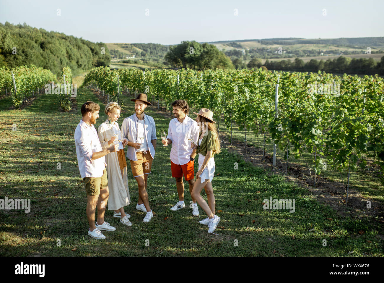 Group of young friends dressed casually hanging out together, tasting wine on the vineyard on a sunny summer morning, beautiful landscape view Stock Photo