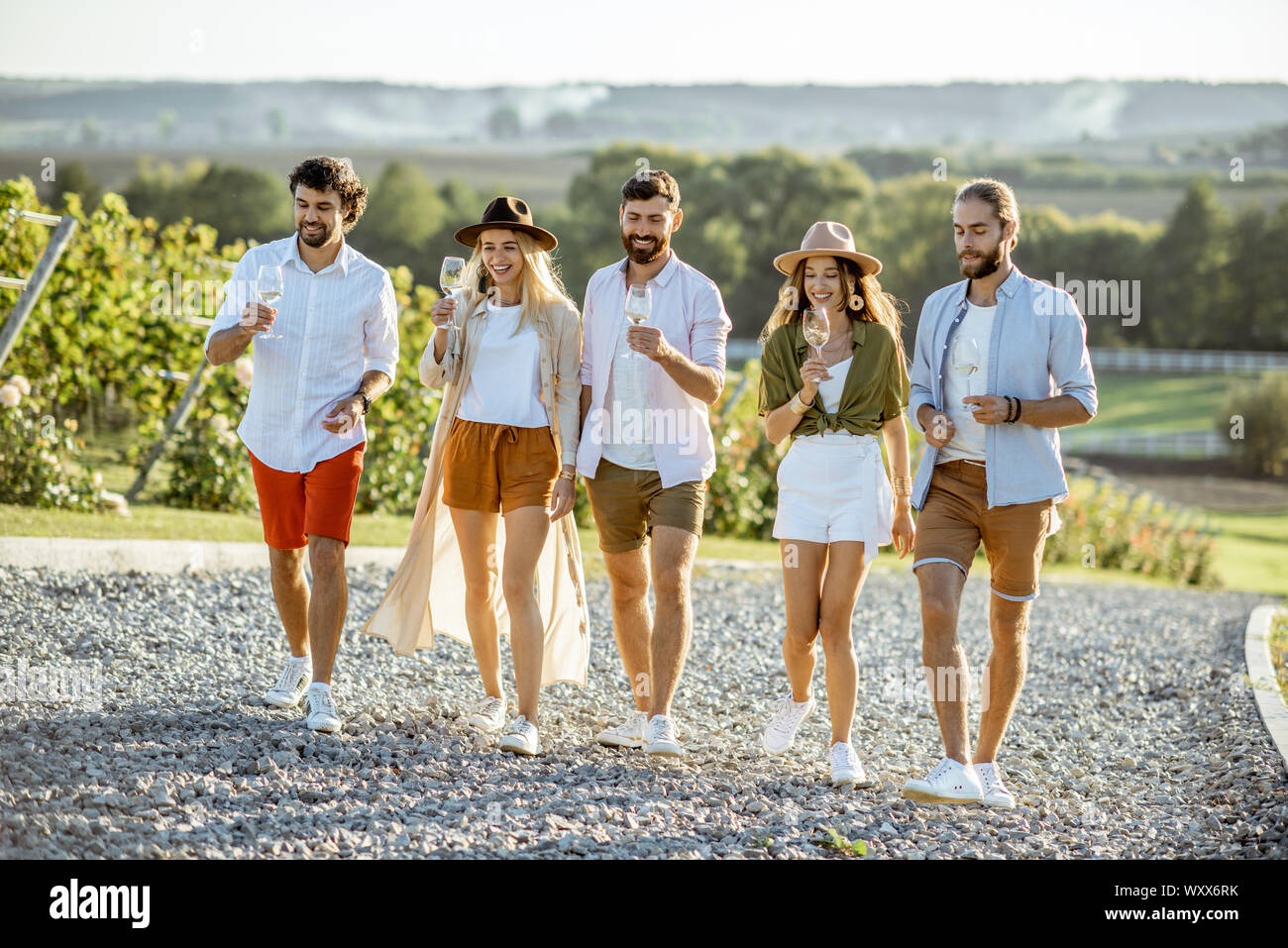 Group of young friends dressed casually hanging out together, walking with wine glasses on the vineyard on a sunny day Stock Photo