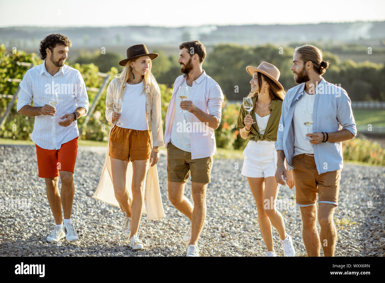 Group of young friends dressed casually hanging out together, walking with wine glasses on the vineyard on a sunny day Stock Photo