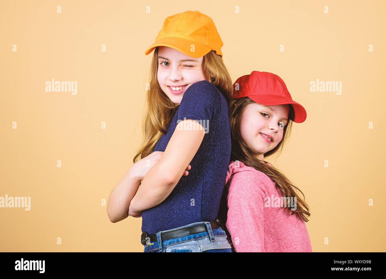Cool girls. Sisters stand back to back beige background. Little cute girls  wearing bright baseball caps. Modern fashion. Hats and caps. Stylish  accessory. Kids fashion. Feeling confident wearing caps Stock Photo -