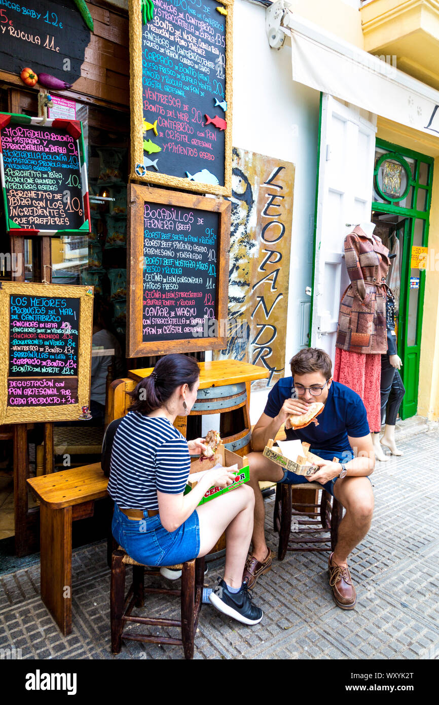 Two people eating sandwiches outside Can Gourmet ,tiny restaurant in Sa Marina offering bocadillos with iberico ham, Ibiza, Spain Stock Photo