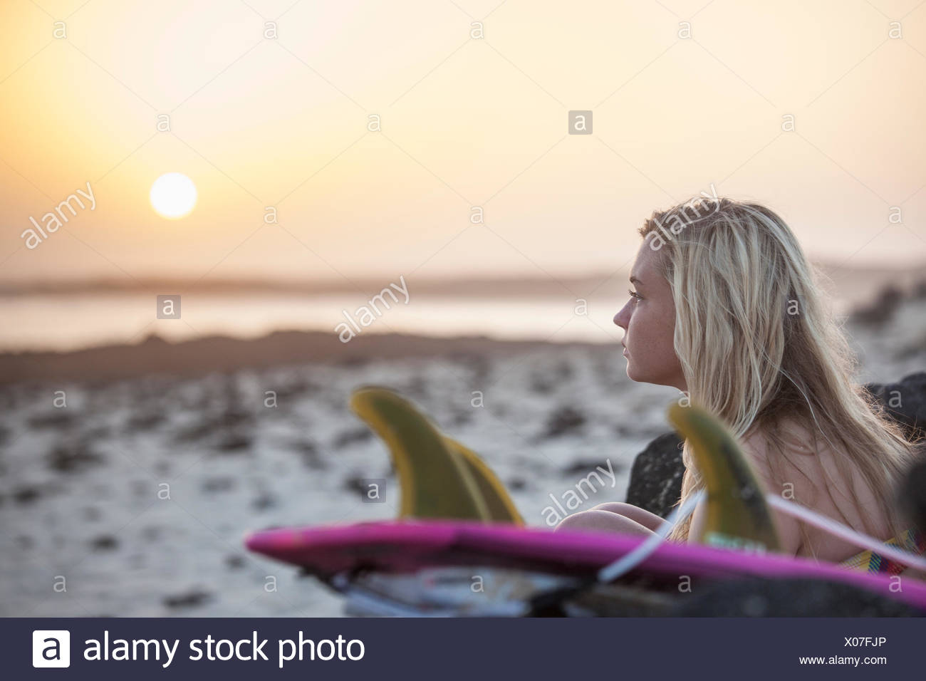 Blonde Surfer Girl Sitting On The Beach At Sunset Stock Photo
