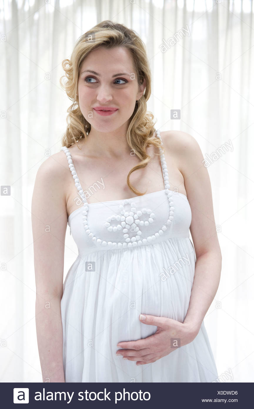 Pregnant Female Wavy Blonde Hair Wearing A White Dress Standing By The Window Hand On Bump Smiling Looking Aside Stock Photo Alamy