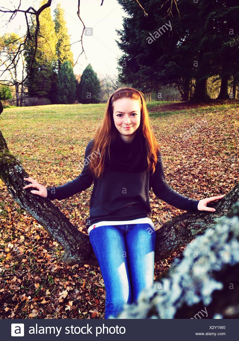 Portrait Of Happy Young Woman Sitting On Branch In Park Stock Photo