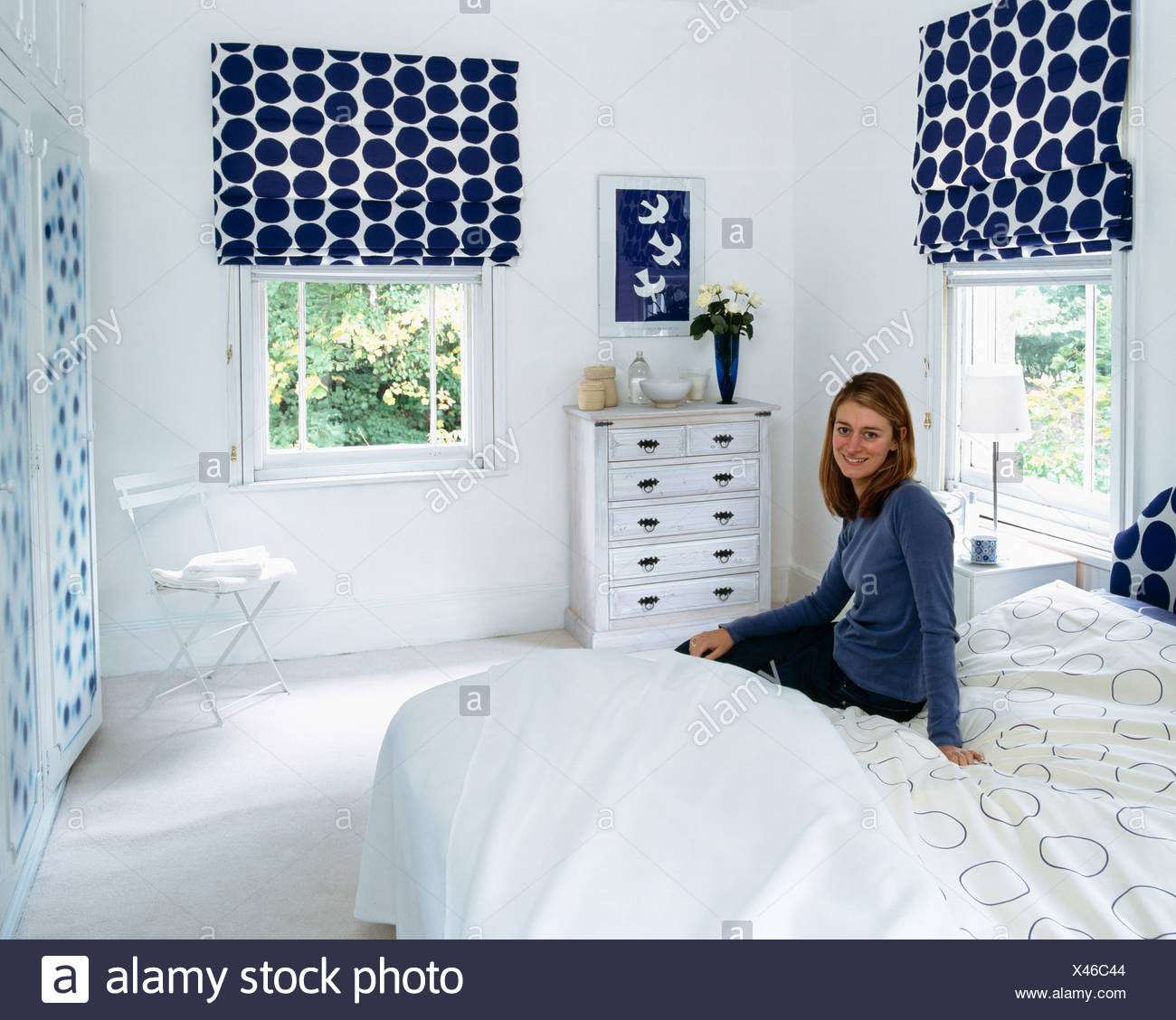 Young Woman Sitting On Bed In White Bedroom With Blue Spotted Blinds On Windows Stock Photo Alamy