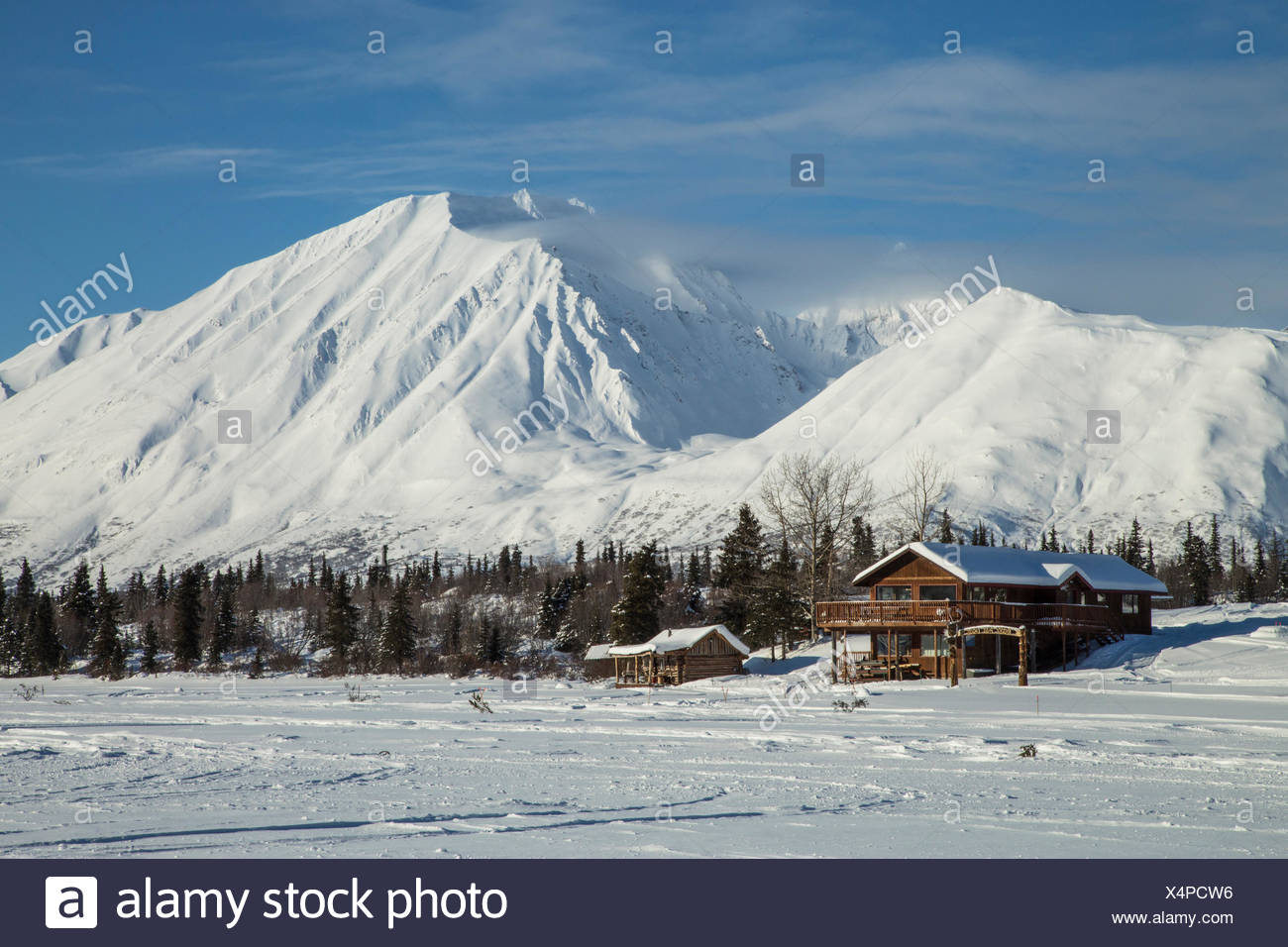 Log Cabins In Alaska Sit At The Base Of A Frozen Lake With Snowy