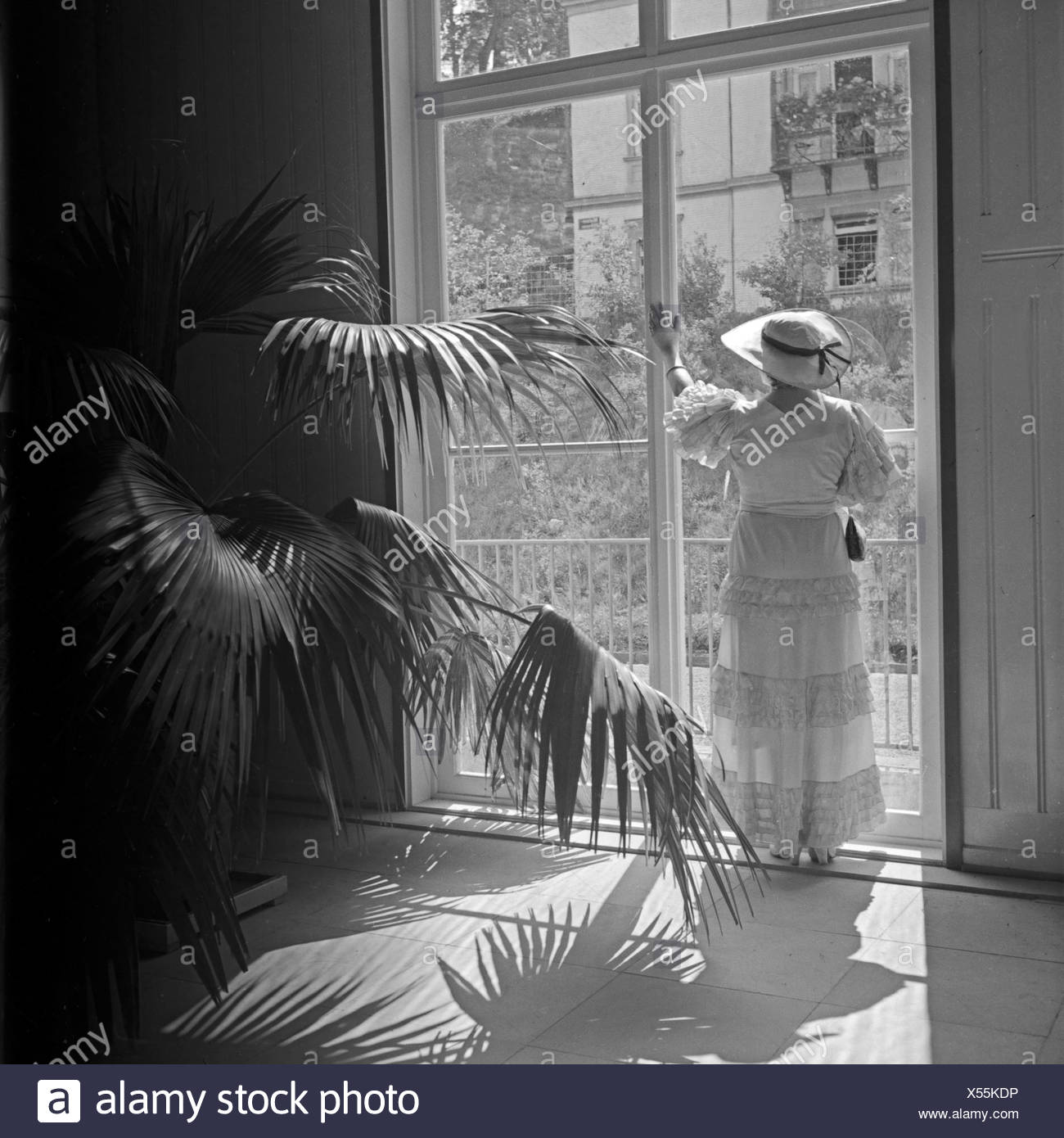 Eine ältere Dame in einem bodenlangen Kleid steht an einem Fenster eines Palmengartens, Deutschland 1930er Jahre. An elder lady in a long dress standing in front of a high window of a palm tree garden, Germany 1930s. Stock Photo