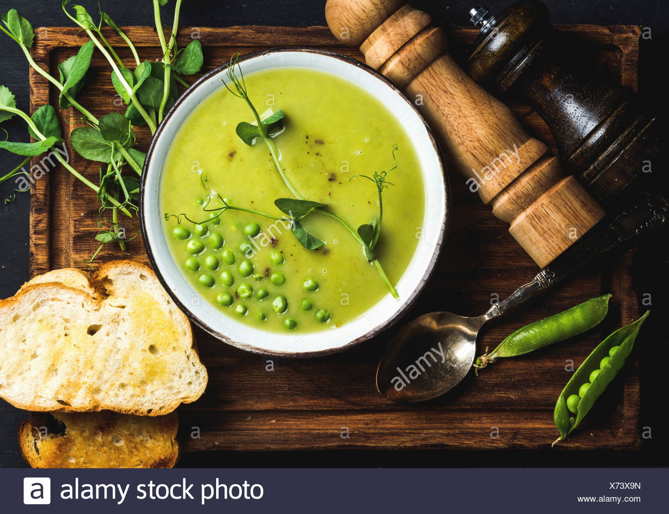 Fresh homemade pea cream soup in white bowl with grilled bread on wooden board over black backdrop, top view, horizontal composi Stock Photo