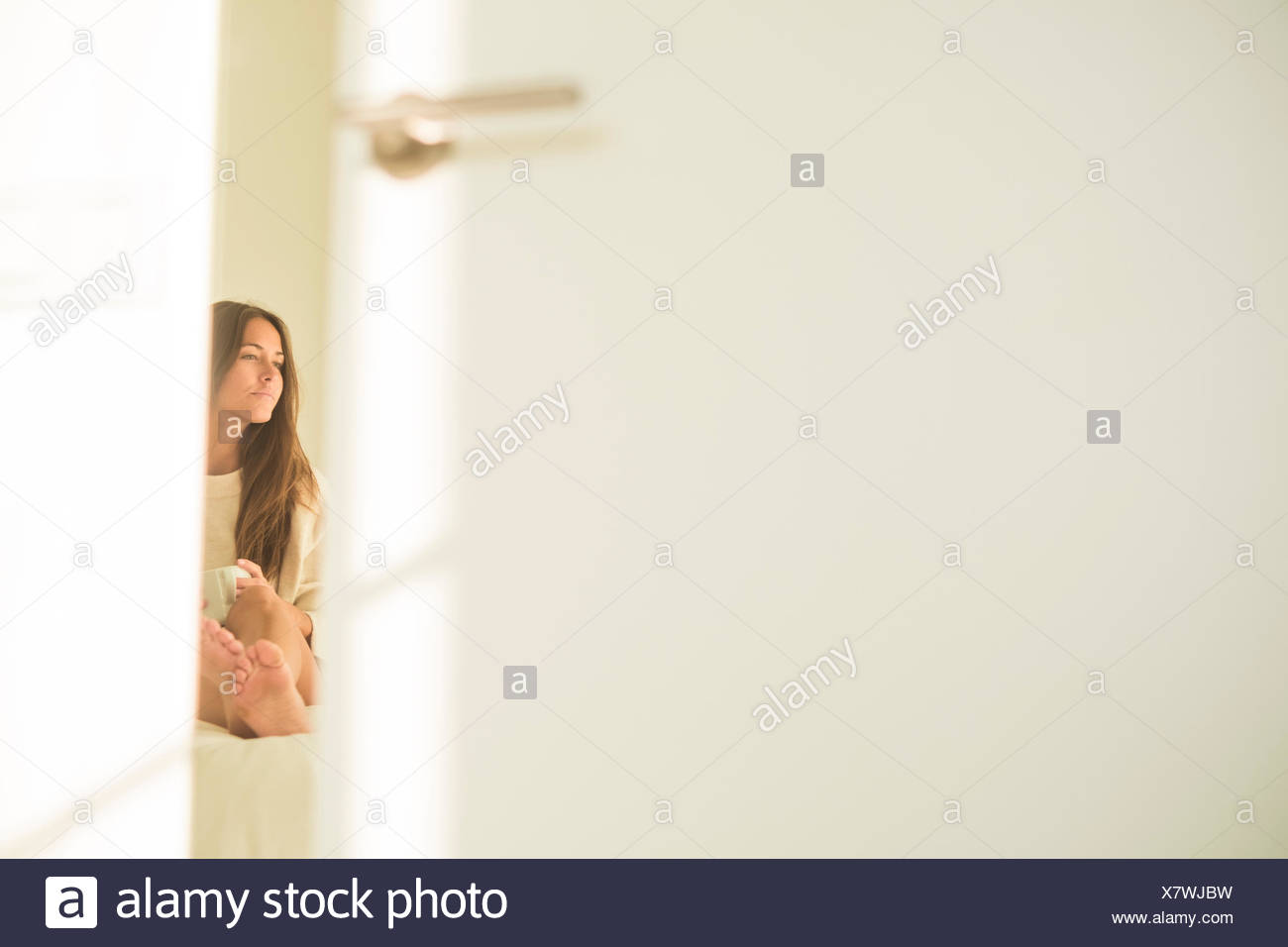 Young Woman Behind Bedroom Door Sitting On Bed With Cup Of